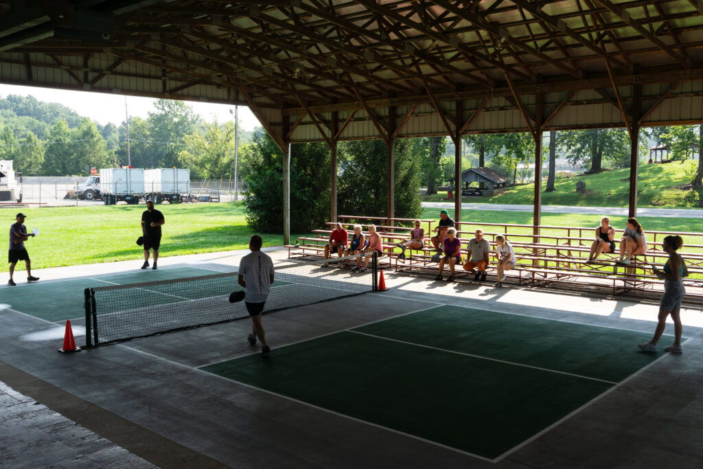 Spectators sitting on benches under a pavilion watching a pickleball match at the Ellettsville event.