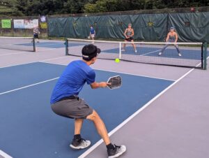 DW Lee hitting a pickleball during a Bloomington Pickleball Club Ladder Event at RCA Park courts, with Susan Cooper and Jodee Davis defending on the opposite side.