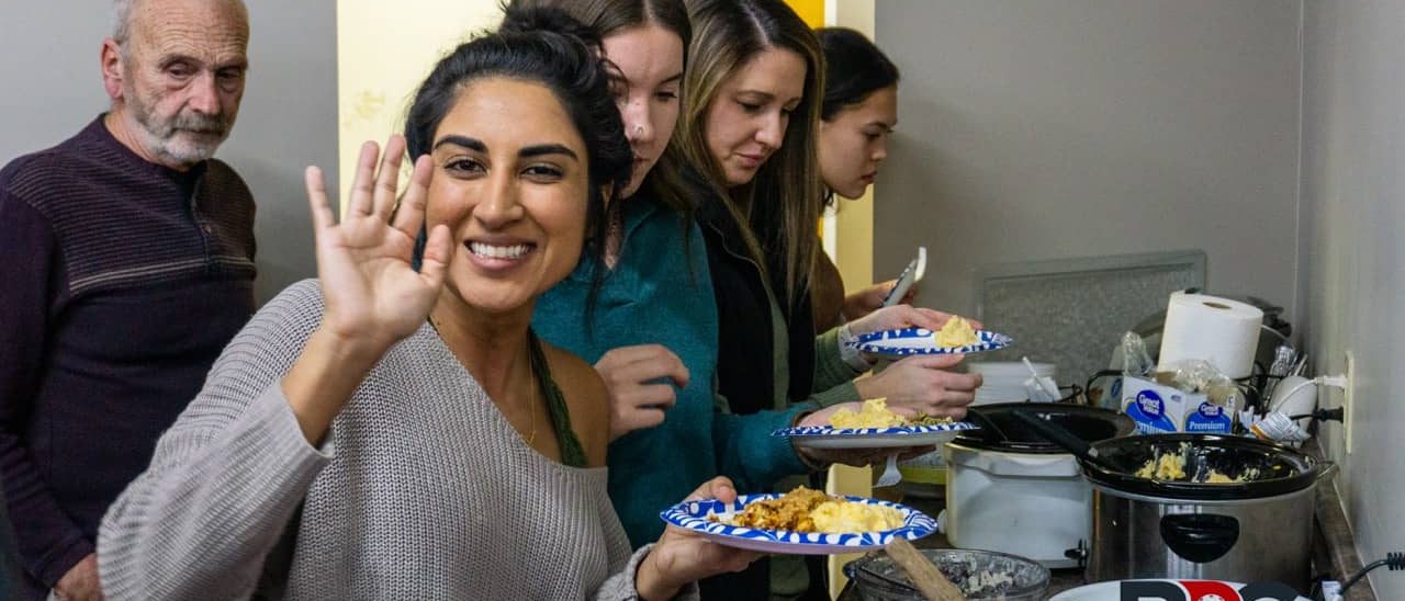 Mehvish Safdar, a Bloomington Pickleball Club member and professional pickleball player, smiling and waving at a social event while holding a plate of food. Other attendees serve themselves from a buffet table in the background.