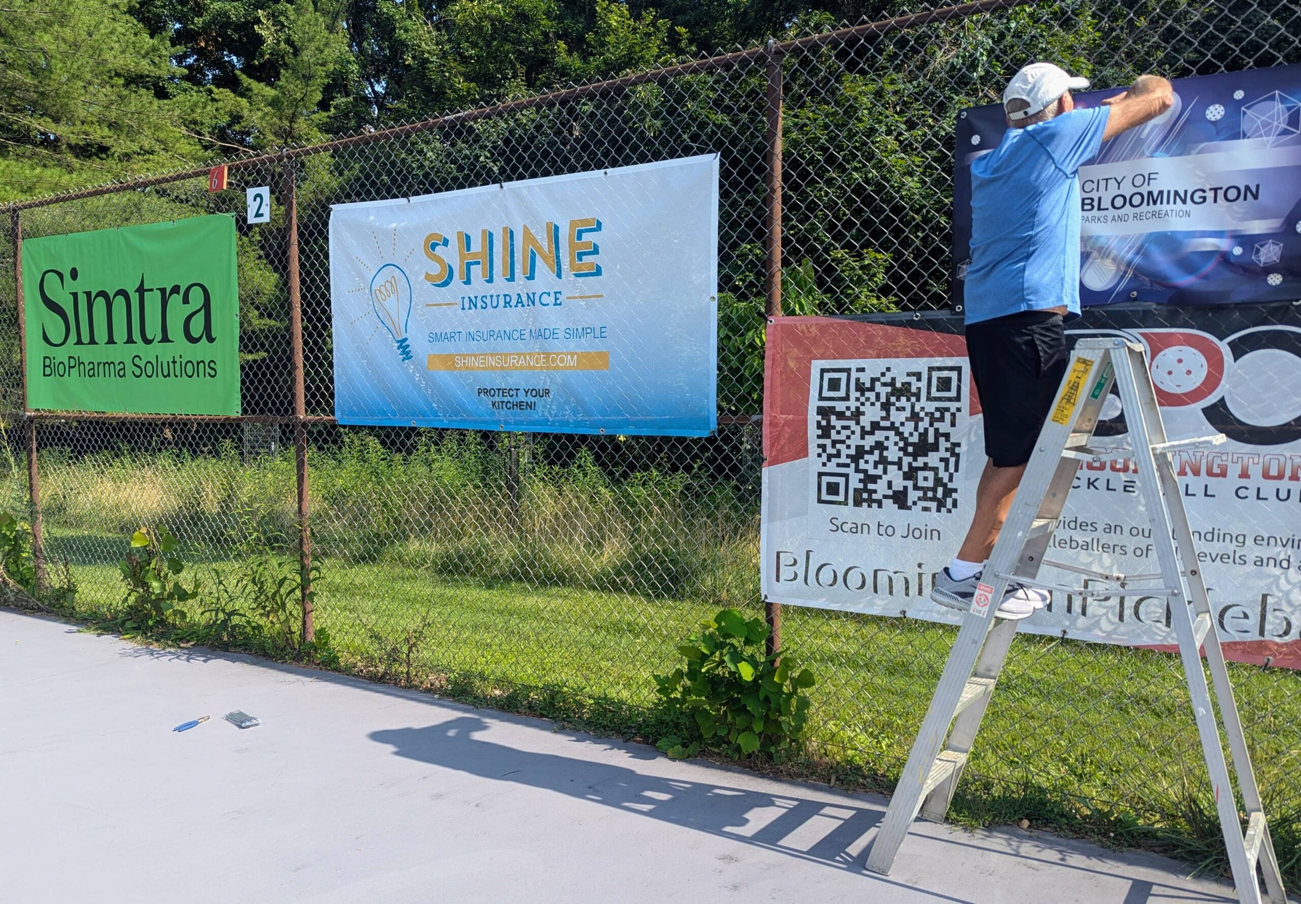 Steve Jackson standing on a ladder hanging sponsor banners for the Bloomington Pickleball Club on a chain-link fence. The banners include advertisements for Simtra BioPharma Solutions, Shine Insurance, and the City of Bloomington Parks and Recreation.