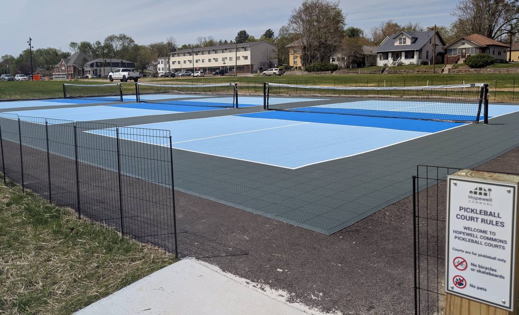 Three outdoor pickleball courts with blue modular tile surfacing and permanent nets, surrounded by a small fence, at Hopewell Commons in Bloomington