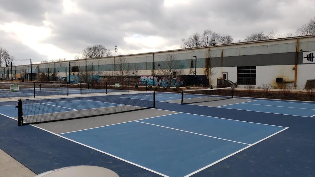 Outdoor pickleball courts at Switchyard Park with blue and gray surfaces, surrounded by fencing and a graffiti-covered industrial building in the background