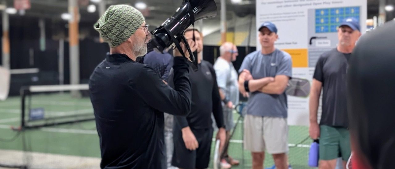 A man in a green knit beanie and black shirt holds a professional camera at an indoor pickleball facility while several club members in athletic wear stand nearby, with a Pickleball Court Use sign visible in the background