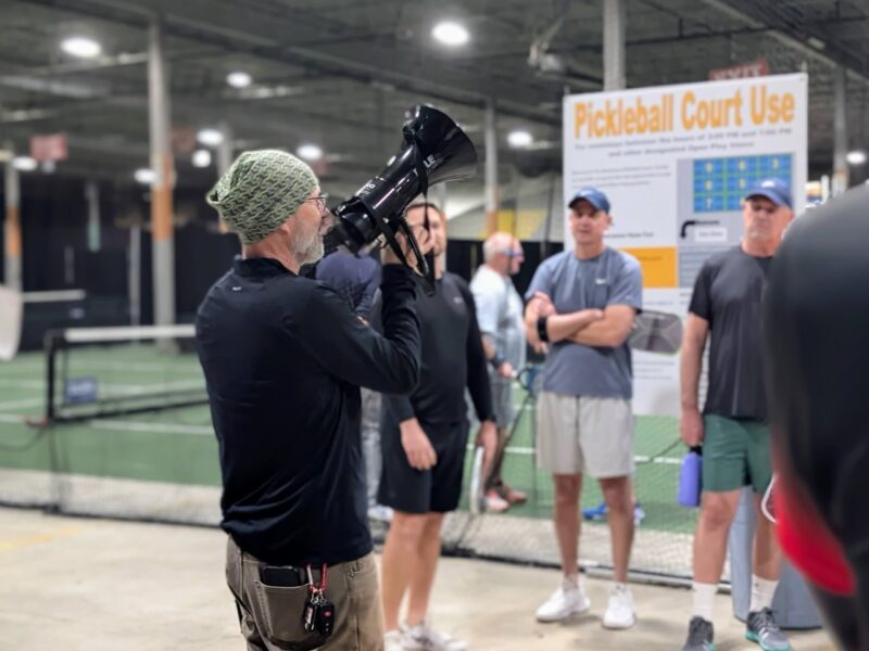 A man in a green knit beanie and black shirt holds a professional camera at an indoor pickleball facility while several club members in athletic wear stand nearby, with a Pickleball Court Use sign visible in the background
