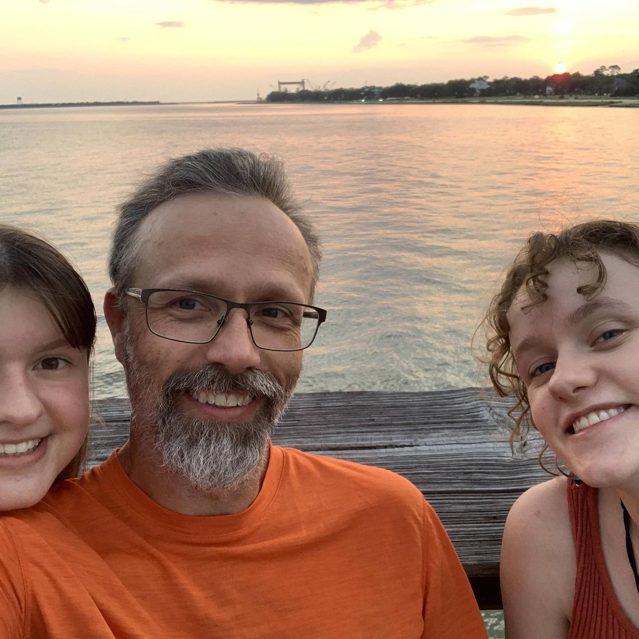 William Bain, pickleball coach and BPC board member, smiles with two young women at sunset on a dock overlooking calm water, all dressed in summer clothes