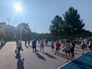 A large group of people gathered on a pickleball court under bright sunlight, preparing for a community event at Bloomington Pickleball Club.