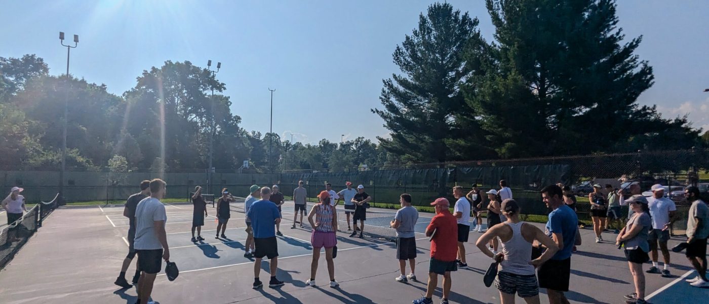A large group of people gathered on a pickleball court under bright sunlight, preparing for a community event at Bloomington Pickleball Club.
