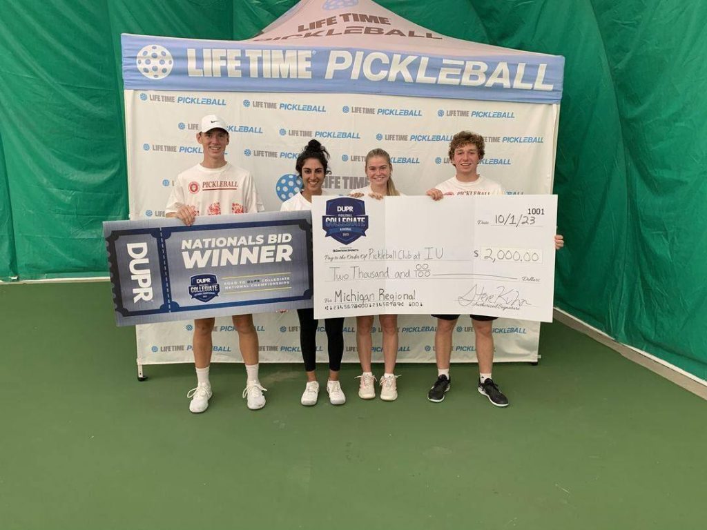 Sam Hutner and Mehvish Safdar, Bloomington Pickleball Club members, stand with their partners holding a Nationals Bid Winner banner and a ,000 check at a Life Time Pickleball event. The group smiles in front of a branded backdrop on an indoor court.