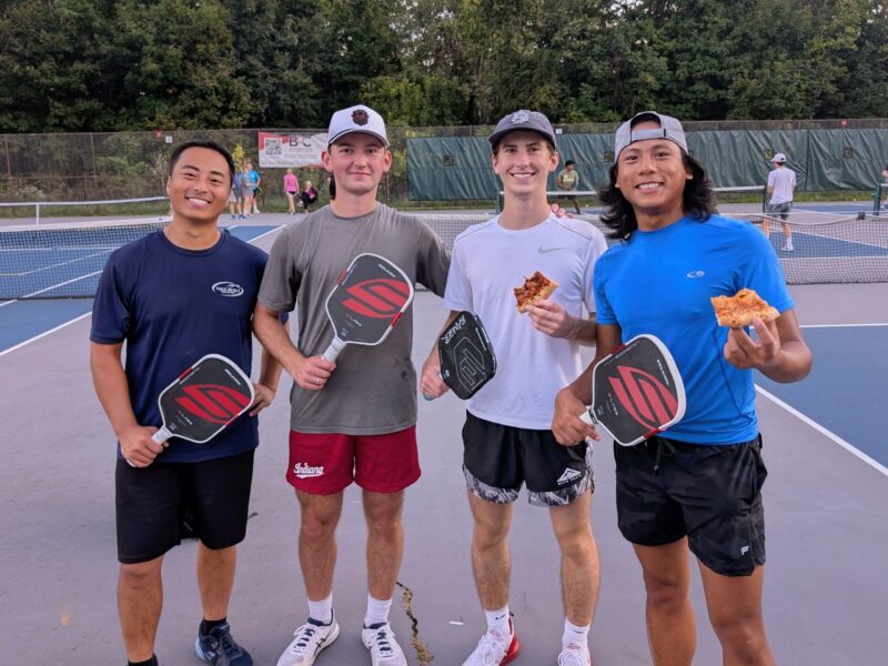 Four smiling pickleball players on court holding Selkirk paddles and pizza slices during a BPC event
