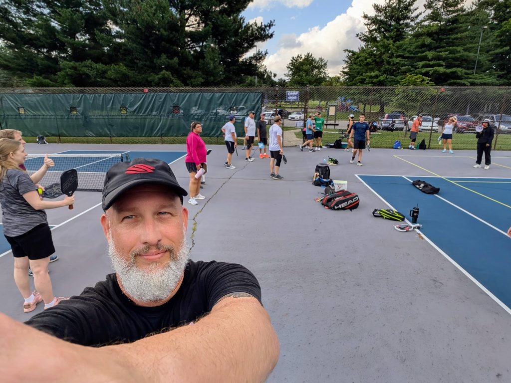 Coach Dave Martin takes a selfie at a busy pickleball court with players warming up and chatting during a Selkirk-sponsored training session