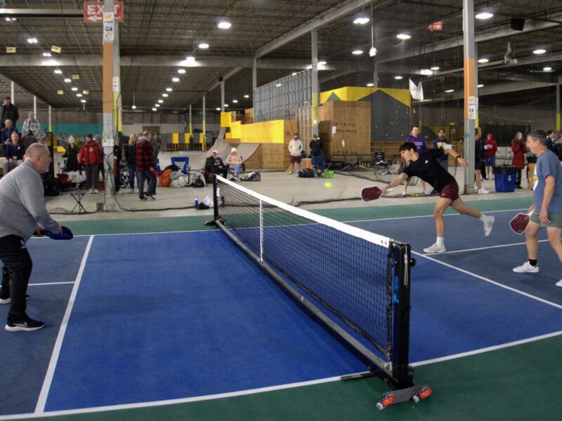 Four players engaging in an intense pickleball match during the Crimson Pickle Grand Opening Tournament in Bloomington, Indiana, January 2024. Spectators watch from the sidelines at the indoor facility.