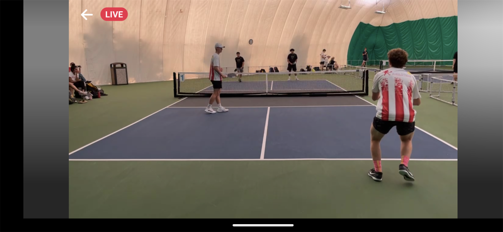 A live pickleball match featuring IU Pickleball team members Carter and Sam. Carter, in a red and white striped shirt, prepares to return a serve while Sam stands ready at the net. The match is held in an indoor facility with green and blue courts and spectators in the background.