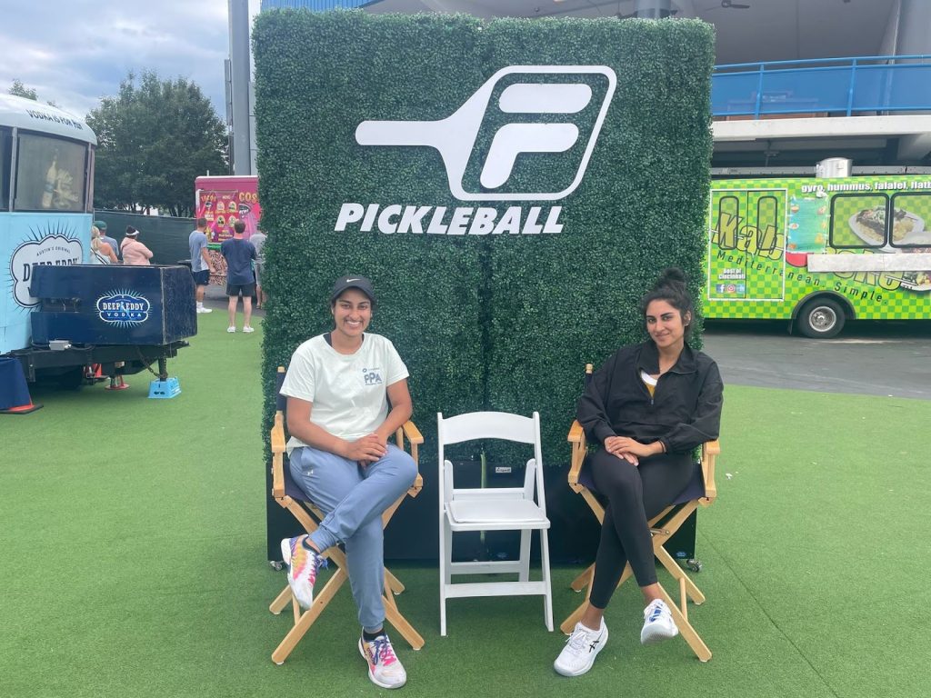 Mehvish Safdar, professional pickleball player, and her sister Komal Safdar sitting in director chairs at a pickleball event. The backdrop features a green wall with the Pickleball logo, and food trucks are visible in the background.