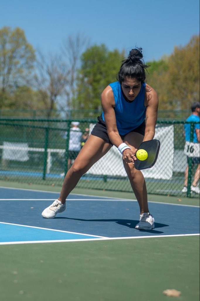 Mehvish Safdar, professional pickleball player, focused during a match as she prepares to hit a low shot with her paddle. She is wearing a blue sleeveless top and black shorts, with a wristband on her right hand. The match takes place on an outdoor court with a green and blue surface.