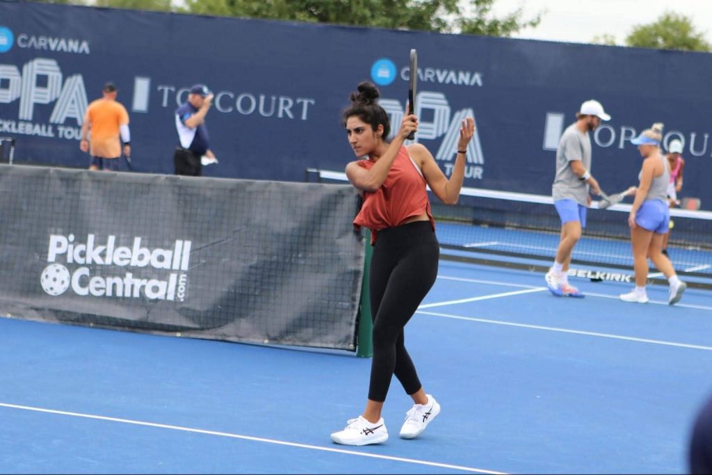 Mehvish Safdar, a professional pickleball player, mid-swing on the court during a competitive match. She is wearing a rust-colored tank top and black leggings, with a determined expression. The backdrop features branding from a professional pickleball tour.