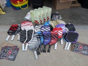 Full display of Selkirk demo pickleball paddles, boxes of balls, banners, and accessories laid out on concrete in front of a garage