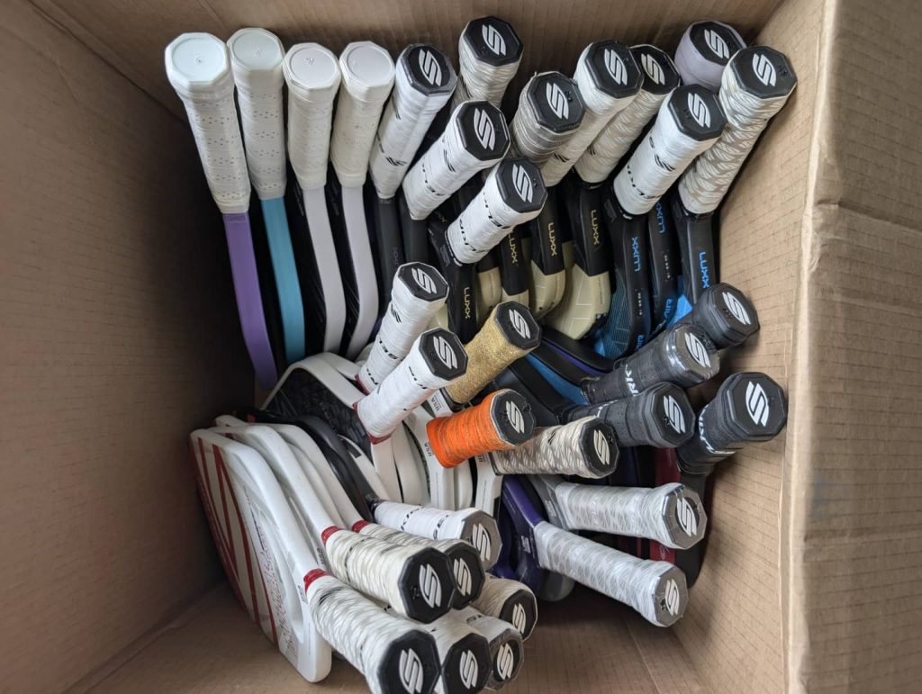 Top-down view of a cardboard box filled with assorted Selkirk pickleball paddles, arranged with handles facing up, showcasing different models and grip colors
