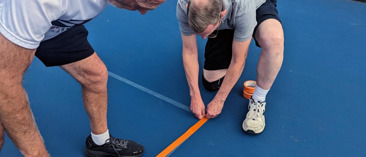 Two volunteers working together to line a pickleball court with tape, preparing it for an event.