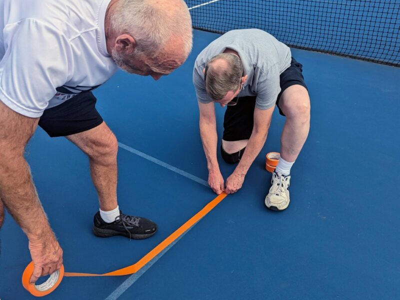 Two volunteers working together to line a pickleball court with tape, preparing it for an event.